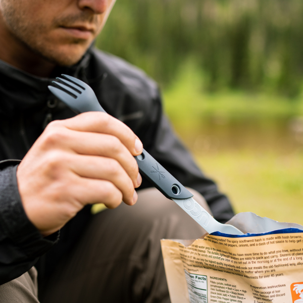 Hand using the fork end of a dark UCO Switch Spork to eat from a foil food pouch outdoors
