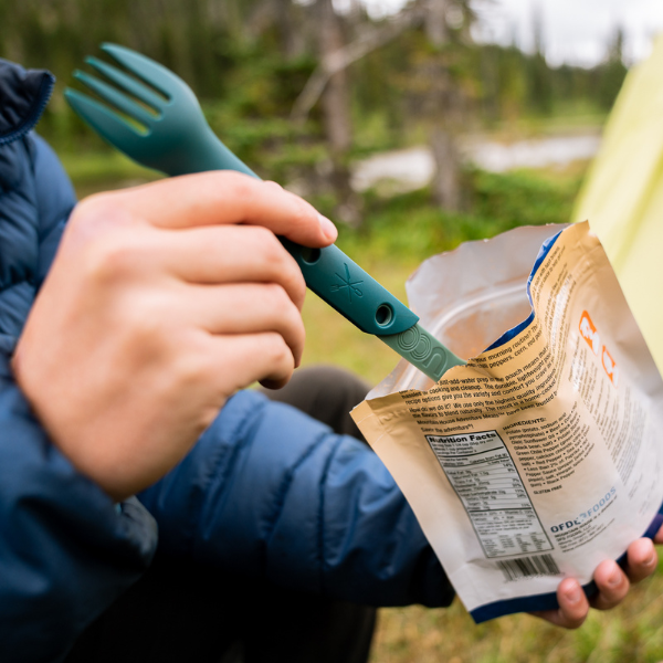Hand using a teal UCO Switch Spork fork end to eat from a foil food pouch outdoors