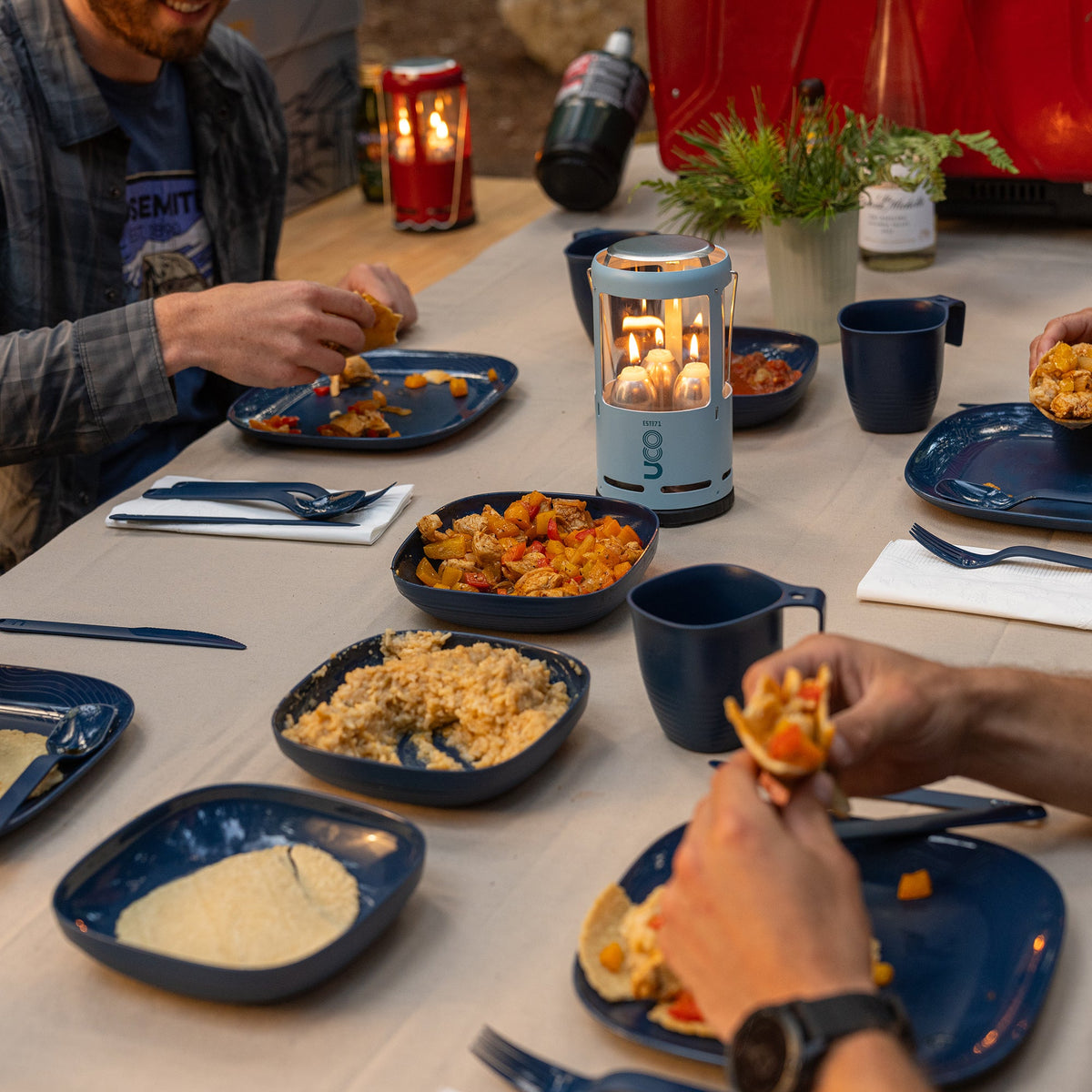 Group of people eating outdoors at a table set with the UCO utensils and a lit candle lantern