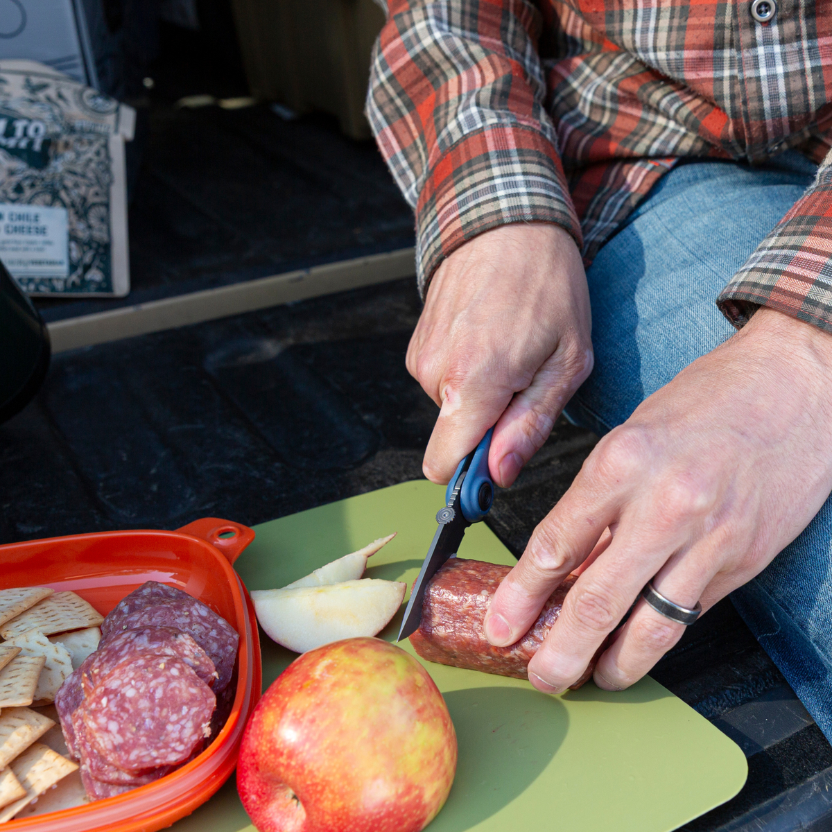 Hand slicing salami and apple on a cutting board with a blue UCO Whidbey Mini folding knife