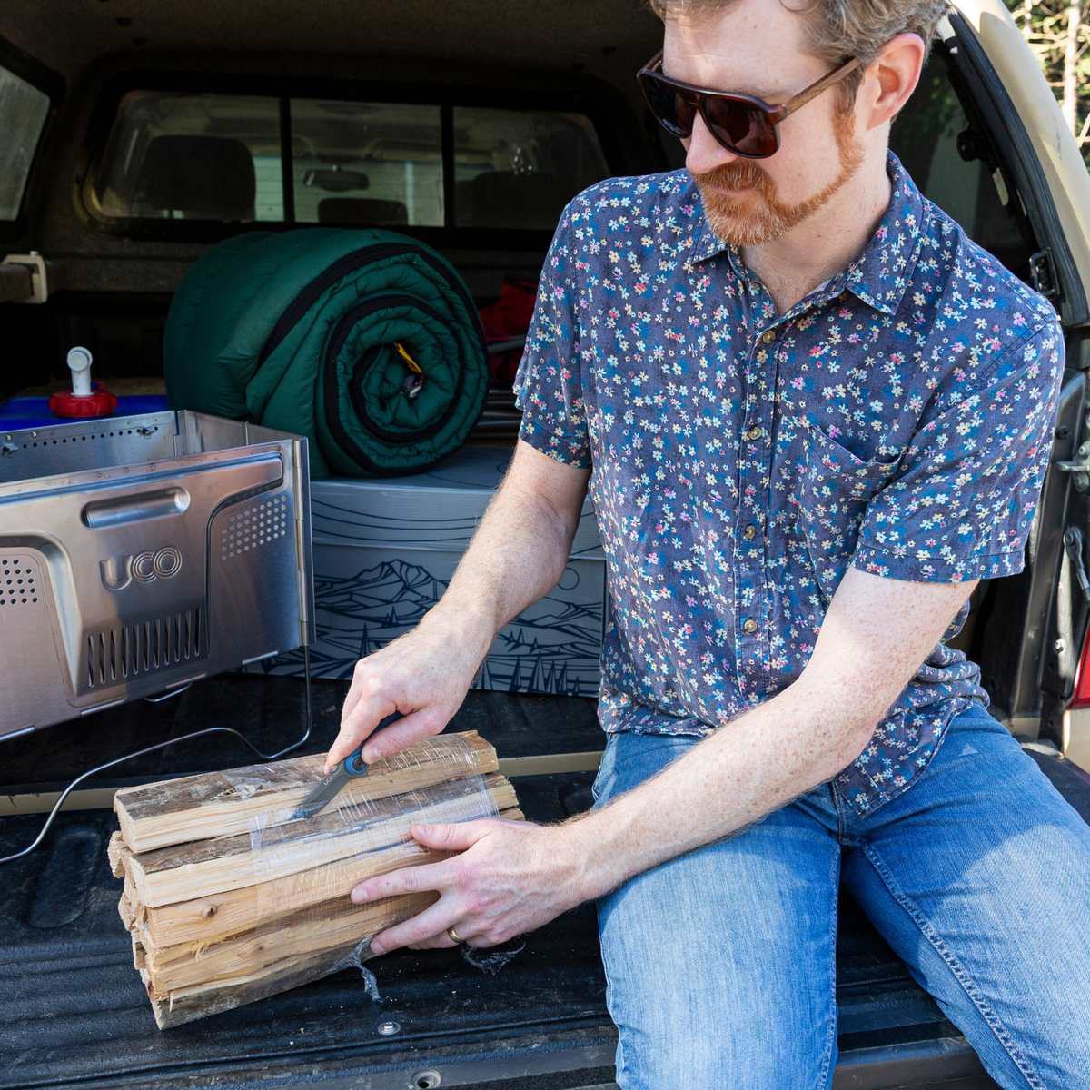 Man in a patterned shirt splitting kindling with a hatchet at a truck tailgate