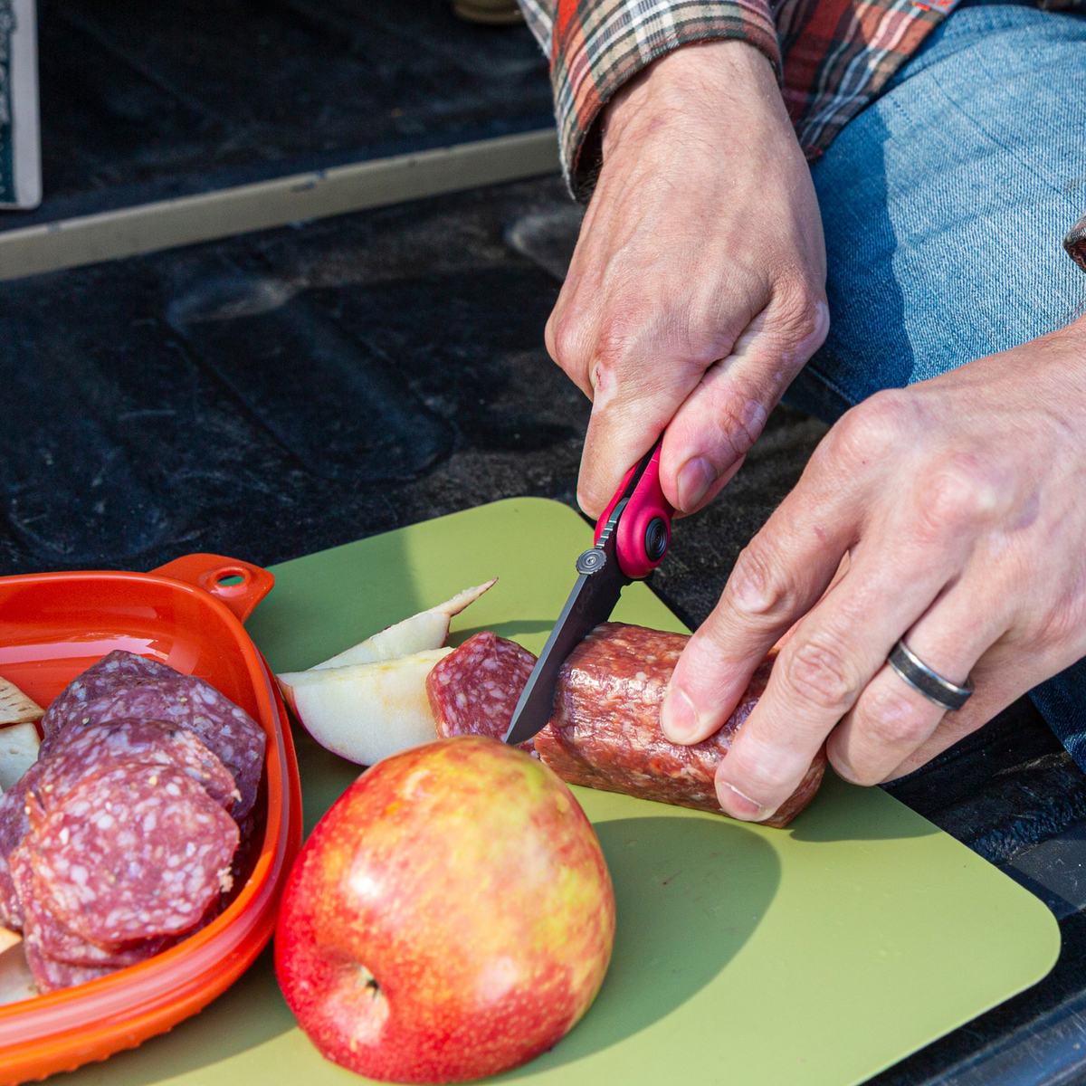 Hands using the red UCO Whidbey Mini folding knife to slice salami and apple on a cutting board