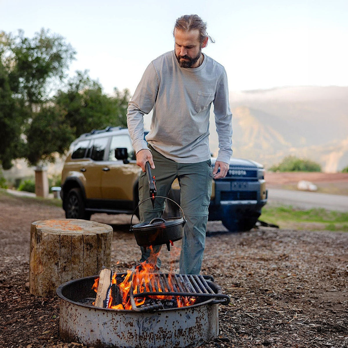 Man at a campsite lifting a cast-iron Dutch oven over a fire ring with a long-handled tool, with a tan SUV parked behind him