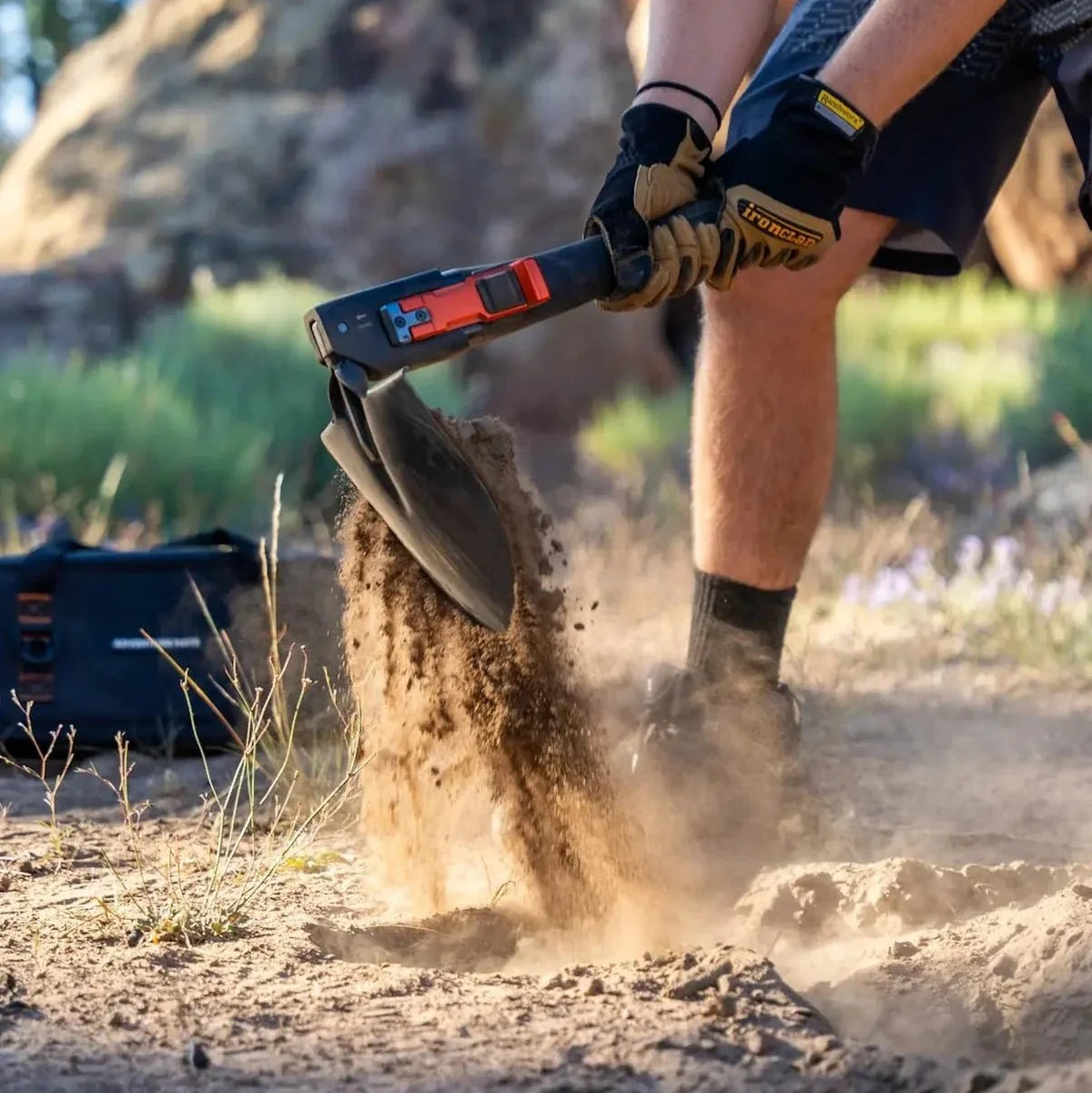 Person in work gloves digging dirt with the Adventure Mate shovel, with a black bag in the background