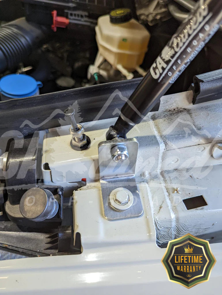 Gas strut attached to a hood bracket in an engine bay, with a lifetime warranty badge in the corner