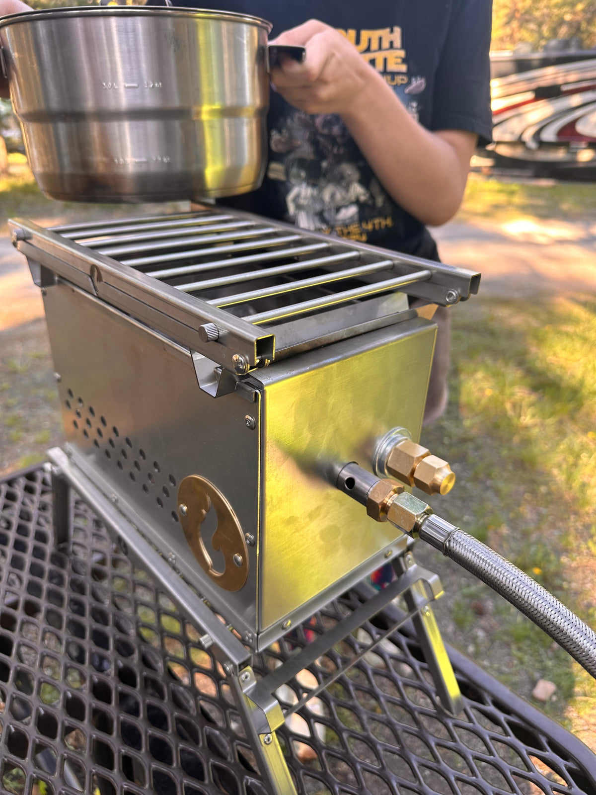 Outdoor photo of a stainless firebox with grill grate on top and a metal cup resting on it, set on a mesh camp table